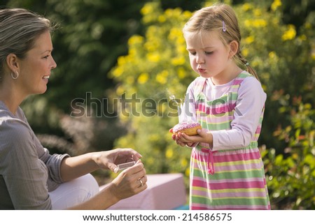 Mother and daughter in garden, daughter blows birthday candles out