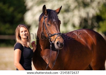 Young woman stands to the right of her horse and smiles at the camera, pictured in landscape format.
