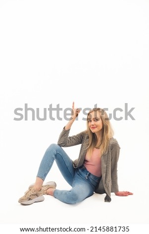 Vertical copy space of a young casually dressed blond-haired girl in her 20s pointing with her index finger up while sitting on the floor. White background. High quality photo