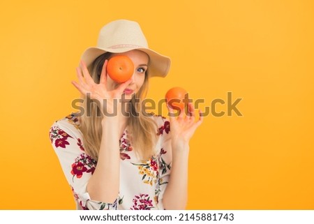 Youthful caucasian blonde girl in her 20s in a hat posing with two oranges or mandarins. Medium studio shot over orange background. High quality photo