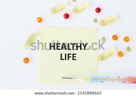 Card with text HEALTHY LIFE on a white background next to a light thermometer and multi-colored tablets, medical concept