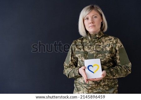 blonde military woman looking at camera while holding card with blue and yellow heart isolated on black