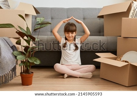 Indoor shot of smiling little girl wearing white t shirt sitting on floor near sofa and cardboard boxes, raised arms and making roof with hands, feels in safety in a new home. Royalty-Free Stock Photo #2145905593