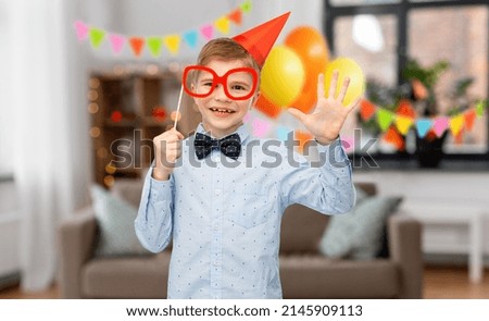 birthday, celebration and childhood concept - portrait of little boy in party hat with glasses over decorated home room background
