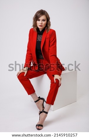 High fashion photo of a beautiful elegant young woman in a pretty red suit, jacket, pants, trousers, black blouse posing on white background. Slim figure, hairstyle, studio shot. Model sits on cube