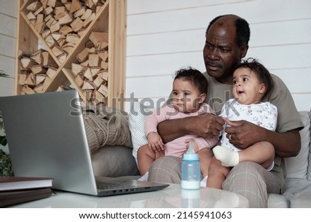 Portrait of modern mature African American father sitting on sofa in living room with twin babies on his lap watching movie in Internet on laptop
