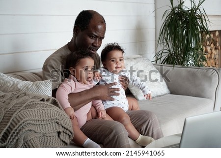 Portrait of mature African American father sitting on sofa in living room with twin baby daughters on his lap watching cartoons in Internet