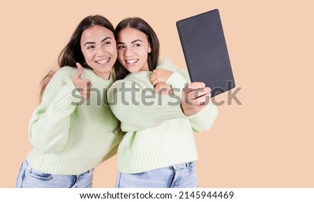 twin sisters taking a picture with a tablet, using a tablet on a beige background, concept of twin sisters of arabian origins