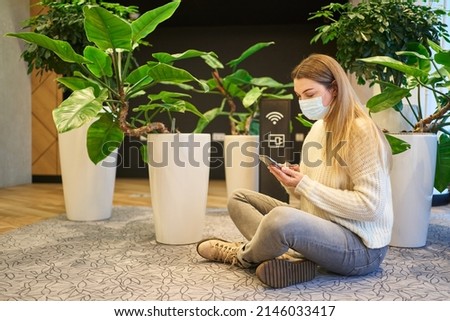 a young woman in a protective mask sits on the floor next to a charging sign and wi-fi and watches something in a smartphone, large house plants are nearby