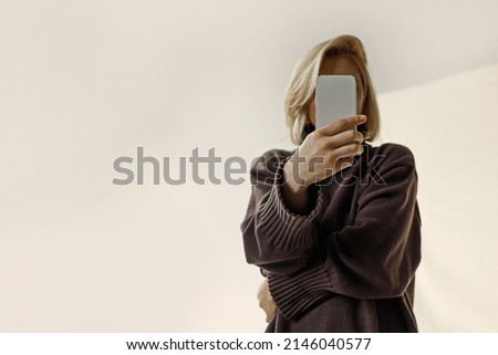 Portrait of a beautiful blonde woman in a brown sweater covering her face with a smartphone, beige background, day Royalty-Free Stock Photo #2146040577