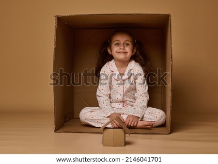 Adorable little European girl sitting inside a cardboard box with a craft house model, smiles looking at camera, posing over beige background, copy ad space. Concept of housing, insurance, real estate Royalty-Free Stock Photo #2146041701