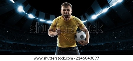 Winner emotions. One professional soccer, football player in football kit standing with ball over dark night stadium with flashlights background. Sport, competition, championship, wow emotion.