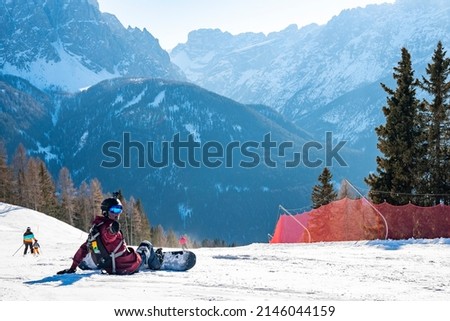 Portrait of snowboarder gesturing thumbs up while sitting on snowy landscape. Scenic view of majestic mountain range against sky. Tourist enjoying winter sport in alpine region.