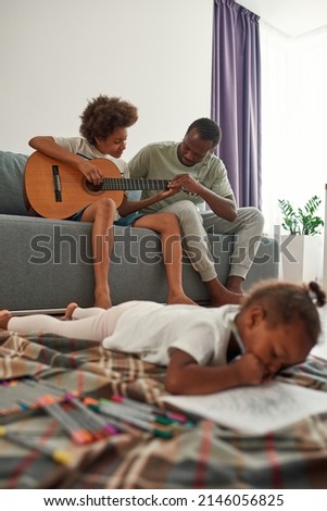 Black father teaching son playing on acoustic guitar on sofa while his little daughter drawing in notebook on blanket on floor at home. Fatherhood and parenting. Domestic entertainment and leisure