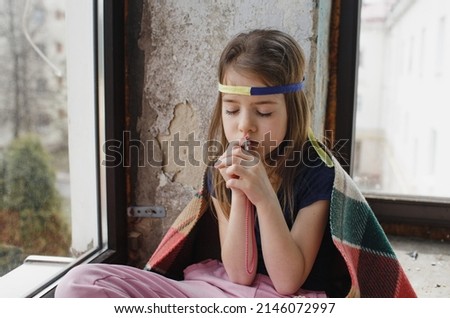 a little Ukrainian girl with a bandage on her head prays for the freedom of Ukraine and holds a cross in her hands. A sad girl is sitting by the window.