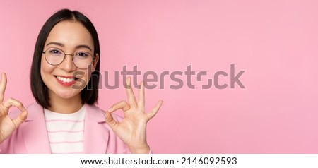 Close up portrait of impressed corporate woman, asian business lady in glasses, showing okay sign, looking amazed at camera, recommending, pink background