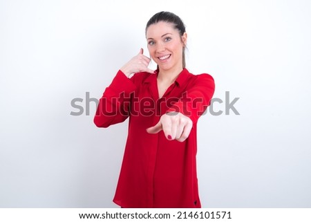 young caucasian girl wearing red shirt over white background smiling cheerfully and pointing to camera while making a call you later gesture, talking on phone