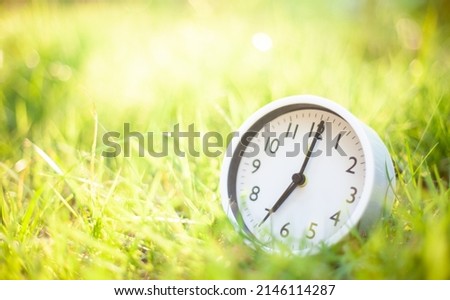 A white clock showing the time placed on a bright green grass. The concept of vacation time. Relaxation. A bright start to a new day.