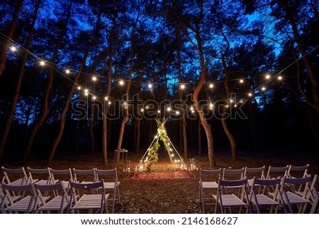 Festive string lights illumination on boho tipi arch decor on outdoor wedding ceremony venue in pine forest at night. Vintage string lights bulb garlands shining above chairs at summer rural wedding. Royalty-Free Stock Photo #2146168627