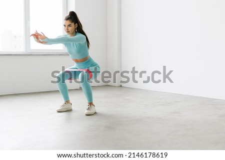 Slim woman works out with resistance band on white wall background indoor. Sporty female athlete in sportswear, doing exercises with elastic, working out for strength and squatting. training at gym. Royalty-Free Stock Photo #2146178619