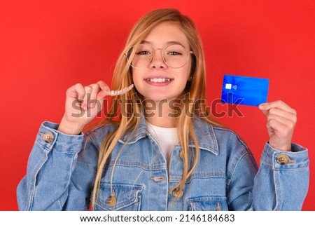 Little caucasian girl wearing glasses and denim jacket standing over red background showing and holding an invisible aligner braces on one hand and a credit card on the other hand. Dental healthcare. 