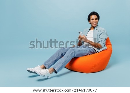 Full size body length fun young black curly man 20s wears white shirt sit in bag chair hold in hand use mobile cell phone typing browsing isolated on plain pastel light blue background studio portrait