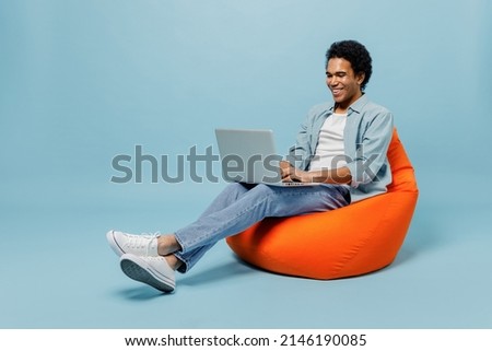 Full size body length young black curly man 20s wears white shirt sit in bag chair hold use work on laptop pc computer typing browsing isolated on plain pastel light blue background studio portrait