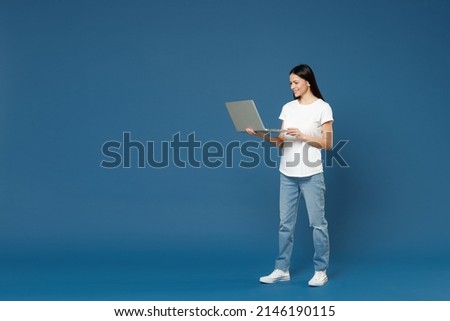 Full length young happy brunette freelancer latin woman 20s in white casual basic t-shirt hold laptop pc computer chat typing sms on social internet isolated on dark blue background studio portrait