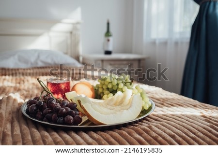 Tray of Fruit Lies on the Bed in Hotel Room, Bottle of Wine Stands on the Background