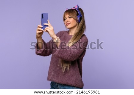 Girl in warm purple oversized sweater and trendy wireless headphones looks at display mobile phone in studio on lilac background. Woman using cell phone to browse social media, listen to podcasts