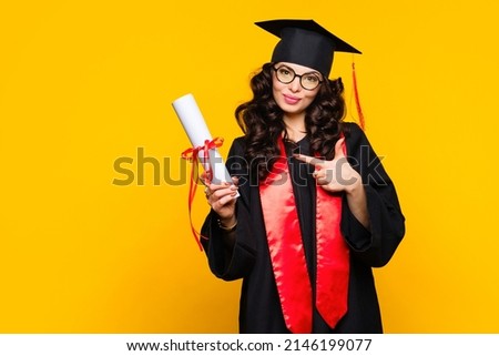 Girl graduate in graduation hat and eyewear with diploma on yellow backdrop. Brunette young woman wearing graduation cap and ceremony robe holding Certificate tied with red ribbon. Education Concept  Royalty-Free Stock Photo #2146199077