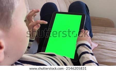 Child lying on the couch and using a digital tablet PC with green screen, back view. Boy holding a tablet with green screen in hand
