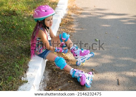 Thoughtful cute girl with Afro-pigtails and wearing sports protective gloves and a helmet sat down to rest on the sidewalk after roller skating. Kid favorite hobby is roller skates. Summer sunny day