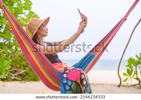 Asian woman sitting on a multicolored cribriform and taking a selfie with a cell phone, During a summer vacation on the beach