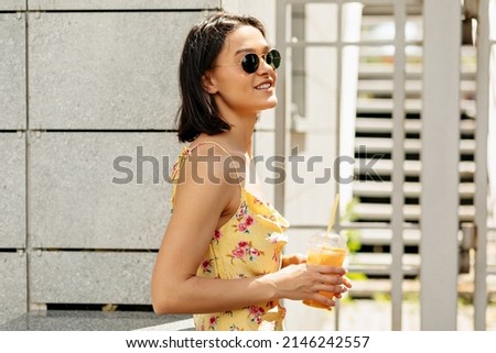 In profile close-up photo of young european woman with happy smile outdoors. Short-haired brunette is holding lemonade, dressed in casual light clothes