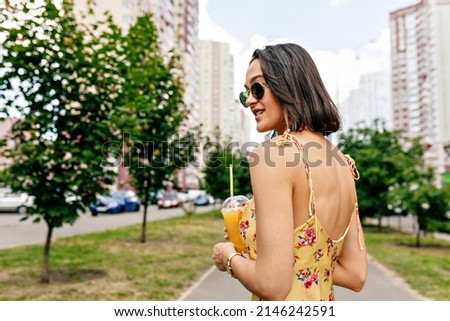 Close up portrait from back of lovely girl with short hairstyle in summer dress and sunglasses walking in the city. Woman is walking with lemonade in summer warm day