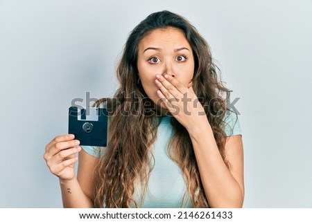 Young hispanic girl holding floppy disk covering mouth with hand, shocked and afraid for mistake. surprised expression 