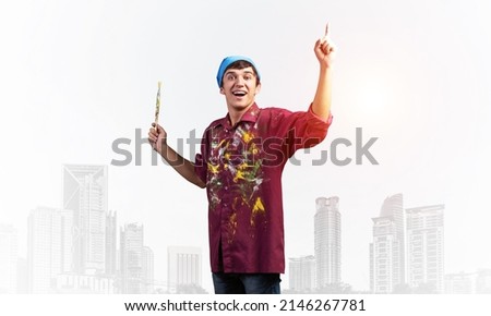 Young emotional artist gesturing with paintbrush. Happy painter in shirt and bandana standing on background modern office buildings. Creative hobby and artistic occupation concept with copy space