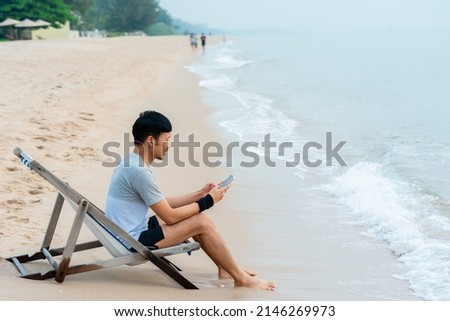 A handsome Asian man, chilling with his mobile phone by the sea in the morning, with cool air and clear skies, comfortably.work for, home