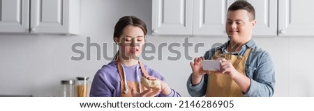 Positive girl with down syndrome holding dough near friend with mobile phone in kitchen, banner