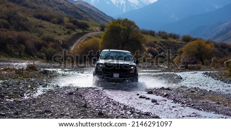 Pick-up truck is off road in the small river in the mountains. View with snow covered mountains in the distance. Landscape of country road with beautiful cloud and sky. Beauty of wild nature. Royalty-Free Stock Photo #2146291097