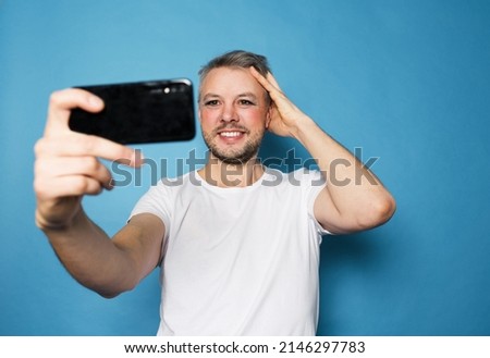 An LGBT man smiles and takes pictures of himself on a smartphone camera standing on an isolated blue background.
