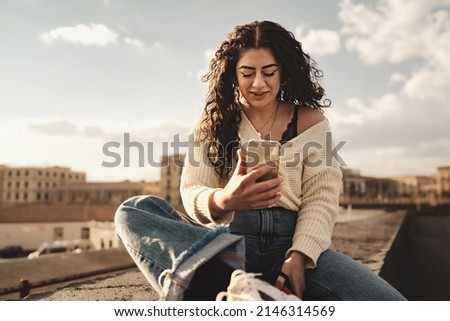 Pretty young teenage curly-haired brunette with nose piercing and dental bracket checking content online with the smartphone, sitting on a low wall by the harbor - urban shot Royalty-Free Stock Photo #2146314569