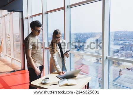 Two colleagues, a man and a woman manager, stand near a table with a laptop in a modern office and discuss a new project. Two business partners in glasses are talking about a new business.