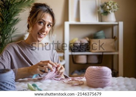 Portrait smiling blonde woman designer knitting clothes and accessories use ribbon yarn and crochet needles. Happy creative female posing at home comfortable interior enjoy art work or hobby Royalty-Free Stock Photo #2146375679