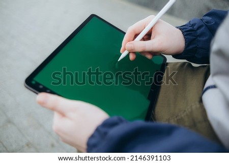 From below of hipster man using modern tablet with stylus while creating new project and sitting on street near glass skyscraper