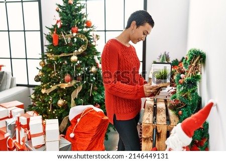 Young hispanic woman smiling confident looking picture standing by christmas tree at home