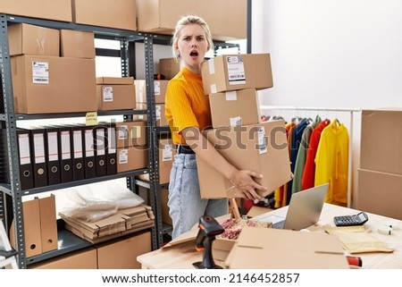 Young blonde woman working at small business ecommerce in shock face, looking skeptical and sarcastic, surprised with open mouth 