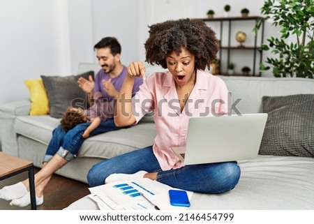 Mother of interracial family working using computer laptop at home pointing down with fingers showing advertisement, surprised face and open mouth 