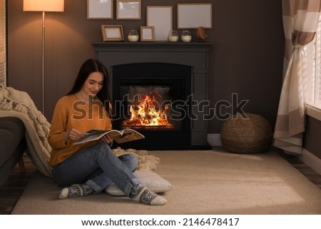 Beautiful young woman reading magazine on floor near fireplace at home. Space for text